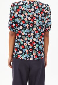 Vista trasera de una persona con una blusa negra con un estampado floral colorido y mangas cortas abullonadas, combinada con pantalones oscuros.
