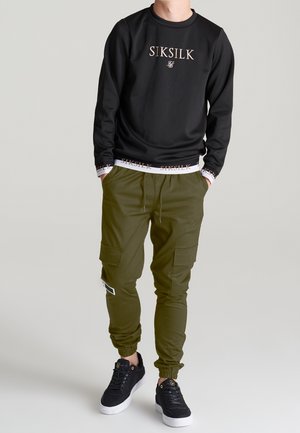 Zwarte sweatshirt met SIKSILK-logo, gecombineerd met olijfgroene cargobroek met zakken en zwarte sneakers met witte zolen.