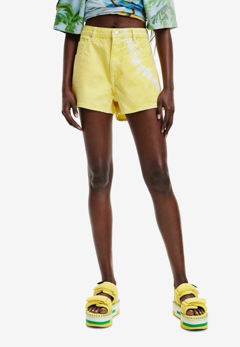 Personne portant un short tie-dye jaune et des sandales plateforme jaunes avec des rayures vertes et blanches, se tenant contre un fond blanc.