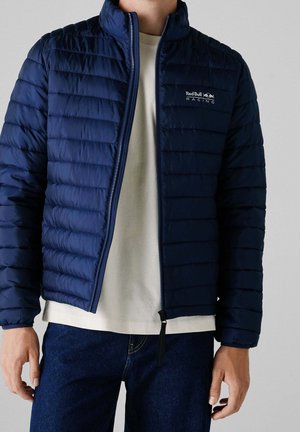 Mężczyzna w granatowej pikowanej kurtce z logo Red Bull Racing, na białej koszuli i ciemnoniebieskich dżinsach, stojący na neutralnym tle.