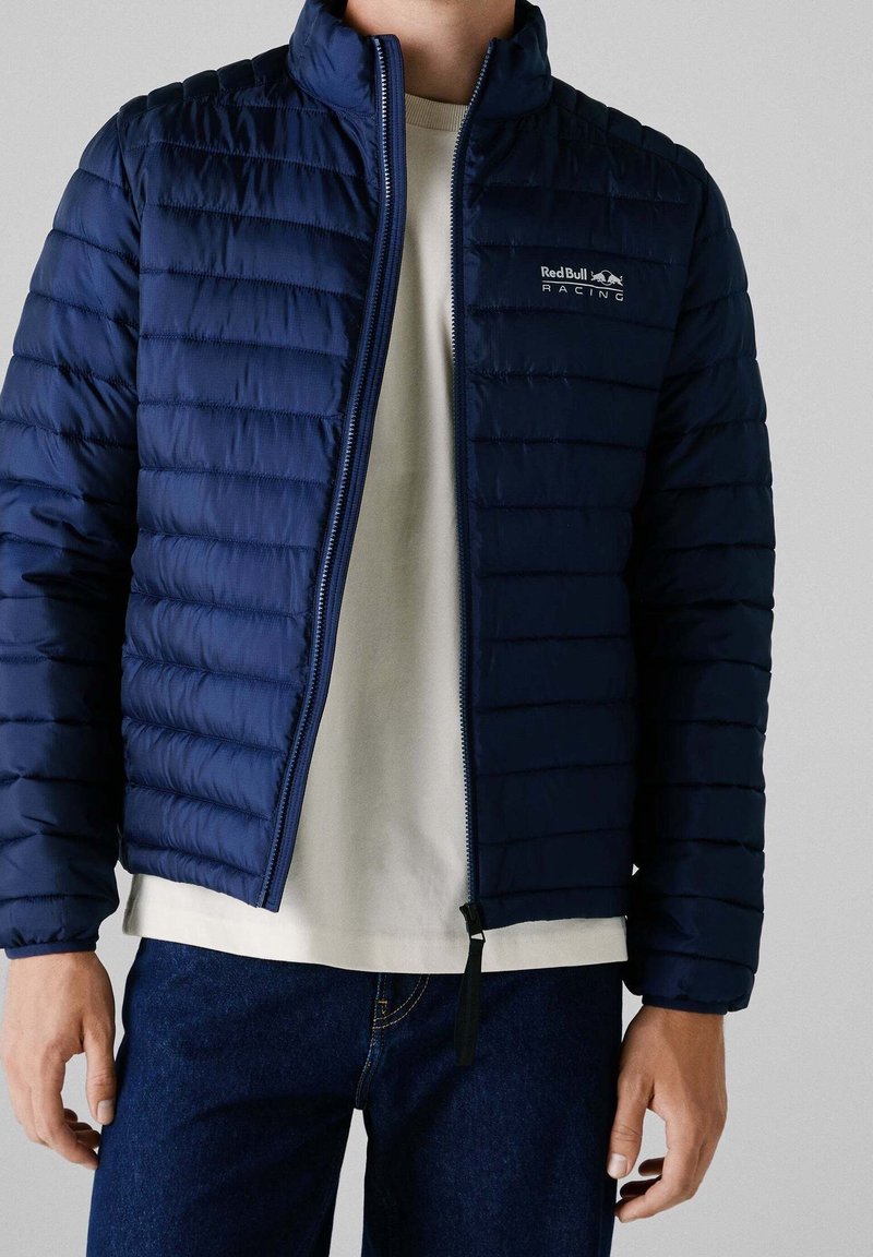 Mann trägt eine marineblaue gesteppte Jacke mit Red Bull Racing-Logo über einem weißen Hemd und dunklen blauen Jeans, steht vor einem neutralen Hintergrund.