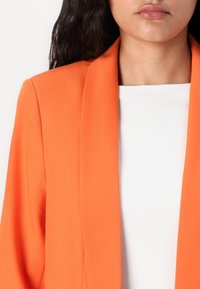 Blazer i livlig orange materiale, med sjalskrave og skræddersyet design, båret over en simpel hvid top. Glat tekstur, tætsiddende silhuet.