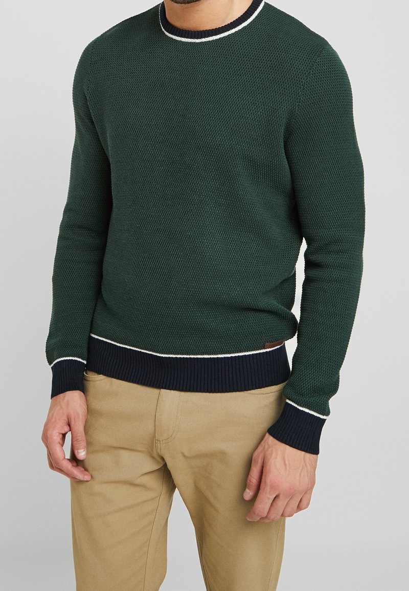 Grön stickad tröja med randiga marinblå muddarna och nederkanten, med en vit detaljrand vid halsringningen. Vävda textur ger djup.