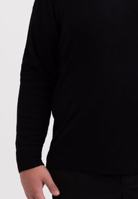 Svart långärmad tröja i mjukt tyg, med rund hals och ribbade ärmar. Texturen verkar jämn och enhetlig genom hela plagget.