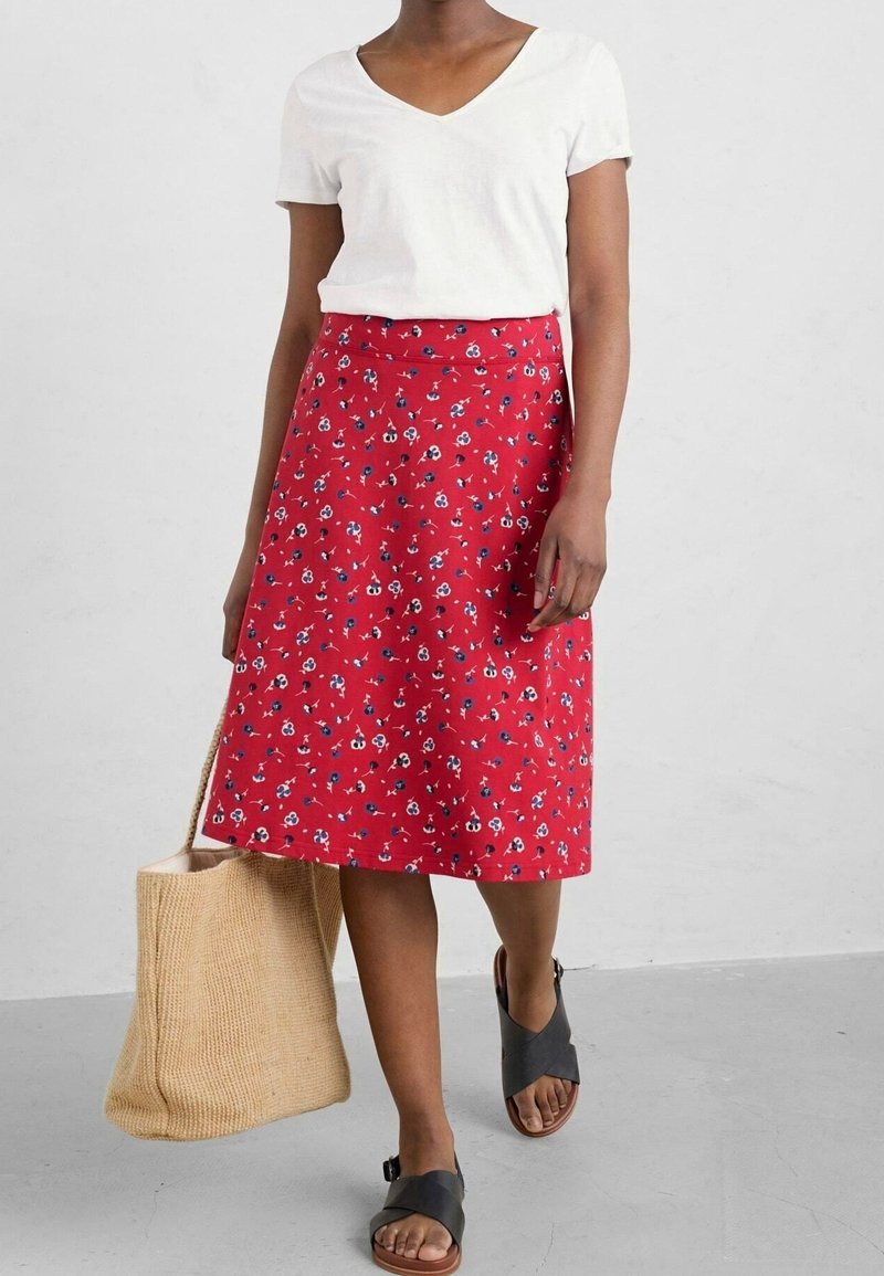 Femme portant un haut blanc à manches courtes en V, une jupe midi fleurie rouge, des sandales noires, et un sac cabas tissé beige.