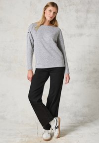 Grauer Pullover mit langen Ärmeln, weitem Halsausschnitt und Knopfdetails an einer Schulter, kombiniert mit schwarzen weiten Hosen und weißen Sneakers.