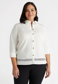 Camicia bianca con maniche a tre quarti, caratterizzata da dettagli in pizzo sulle spalle e un motivo decorativo a intaglio sull'orlo.