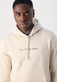 Felpa beige con cappuccio e cordino, caratterizzata dal testo "Marc O'Polo DENIM" ricamato in grigio. Morbido al tatto, con tasca a marsupio sul davanti.