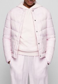 Giacca a vento rosa chiaro con chiusura frontale a bottoni a pressione, caratterizzata da un design trapuntato e tasche laterali, indossata sopra un maglione abbinato.