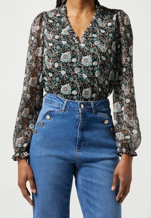 Vrouw draagt een zwarte, met bloemen bedrukt blouse met lange mouwen van doorschijnende stof en hooggetailleerde blauwe spijkerbroek met knoopdetails aan de zijkanten.