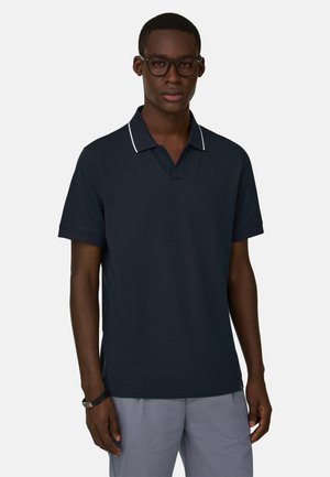 Mann mit schwarzer Brille und navyblauem Poloshirt mit weißer Einfassung, kombiniert mit hellgrauer Hose, vor neutralem Hintergrund stehend.