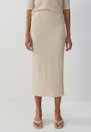 Kvinde iført en tekstureret beige midinederdel med en matchende top og beige sandaler med åbne tæer, stående mod en ensfarvet lys baggrund.