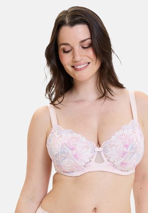 Mujer sonriendo y mirando hacia abajo, vistiendo un sujetador de encaje rosa claro con patrones florales sobre un fondo liso.