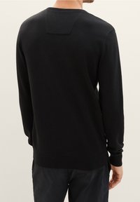 Maglione nero lavorato a maglia con maniche lunghe, scollo rotondo e piccolo dettaglio a toppa sulla parte superiore della schiena, caratterizzato da una texture liscia.
