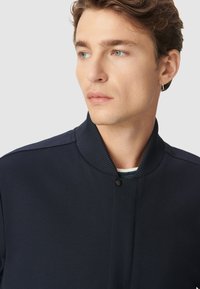 Chaqueta bomber marina con cuello acanalado y cierre de botón a presión, presenta una textura suave y un diseño minimalista.