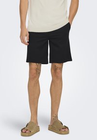 Sorte shorts i en afslappet pasform, lavet af et glat stof. Sammen med brune slip-on sandaler. Synlige tatoveringer på benene.