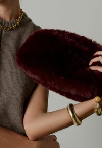 Bordeaux nep-vacht clutch met een zachte textuur, met een glad oppervlak. Gecoördineerd met een grijze trui en een kettinghalsband in goudkleur.