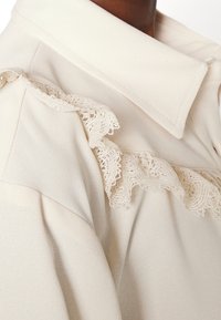 Cream-colored garment with a wide collar and delicate lace trim detail along the shoulder and chest area.