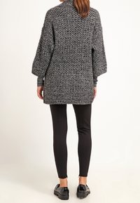 Cardigan grigio oversize lavorato a maglia con texture a coste, spalle cadute e taglio dritto, abbinato a leggings neri e scarpe.