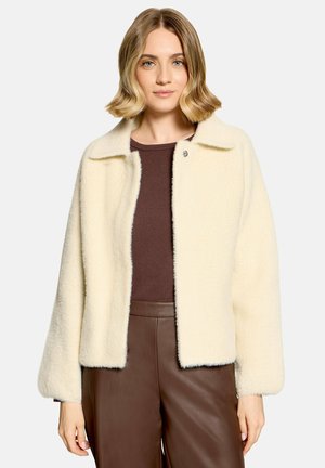 Femme aux cheveux blonds mi-longs portant une veste crème pelucheuse par-dessus un haut marron et un pantalon en cuir marron, debout devant un fond uni.