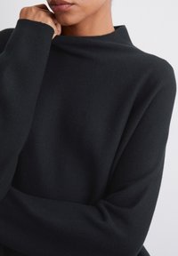 Pull en maille noire avec un col haut et replié et des manches larges. Le tissu semble doux et texturé, mettant en valeur une coupe décontractée.