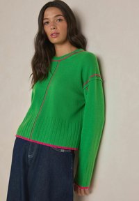 Vihreä neulottu villapaita, jossa on resorit helmassa ja hihansuissa. Kaula-aukon ja sauman kohdat on korostettu vaaleanpunaisilla yksityiskohdilla. Päällä tummat farkut.