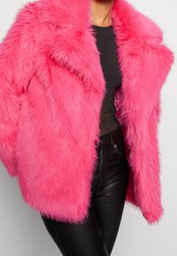 Jasnoróżowy sztuczny futerko z szerokim kołnierzem i długimi rękawami. Charakteryzuje się puszystą fakturą i luźnym krojem, w zestawieniu z czarnymi dopasowanymi spodniami.