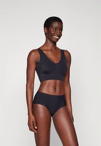 Smilende kvinne med kort hår iført en svart sømløs crop top og matchende svarte sømløse truser, stående mot en ensfarget bakgrunn.