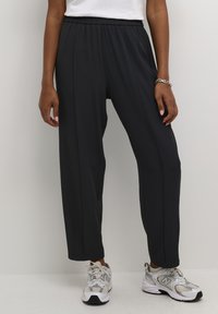 Pantalons de jogging noirs à taille élastique avec une coupe droite et des détails sur les coutures latérales, associés à des chaussures de sport grises et blanches.