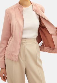 Jasnoróżowa kurtka o miękkiej, teksturowanej fakturze, z zamkiem błyskawicznym i wewnętrzną kieszenią, zestawiona z białym prążkowanym topem i beżowymi spodniami.