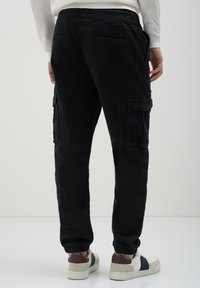 Mann in schwarzen Cargohosen mit elastischen Bündchen und weißen Sneakers mit dunklen Akzenten, der vor einem schlichten hellen Hintergrund steht.