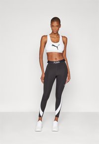 Sorte leggings med hvite detaljer og PUMA-logo, kombinert med en hvit trenings-BH med svart logo, begge laget av elastisk stoff.