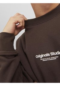 Personne tirant sur le col d'un sweat-shirt marron avec un texte blanc indiquant « Originals Studio, brought to you by Jack & Jones, designed in Copenhagen. »