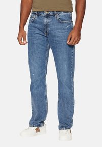 Jeans in denim blu con taglio dritto, caratterizzati da lievi rotture, cinque tasche e chiusura a bottoni. Indossati con sneaker bianche.