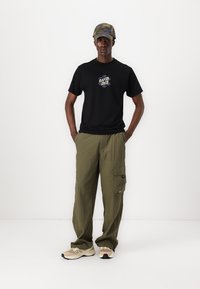 Tricou negru cu imprimeu grafic, pantaloni cargo verzi măslinii cu buzunare laterale, adidași bej și crem și o șapcă de camuflaj.
