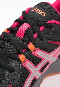 Chaussure de sport noire avec des accents roses et oranges. Elle présente un dessus en tissu texturé, des lacets contrastants et un logo réfléchissant.