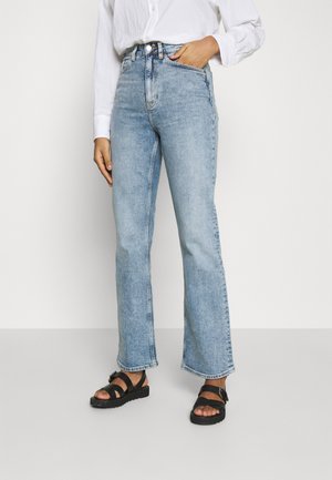 Frau trägt hellblaue Jeans mit geradem Bein, weiße Bluse mit langen Ärmeln und schwarze Sandalen, steht mit einer Hand in der Tasche.