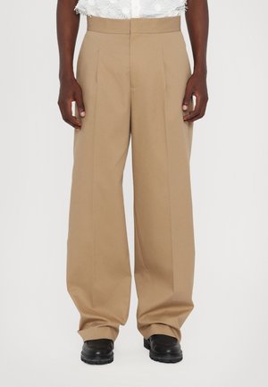 Personne portant un pantalon beige taille haute à jambes larges et des chaussures noires, debout devant un fond blanc uni.