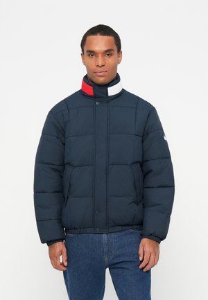 Jachetă puffer marină, cu guler înalt, prezentând un accent roșu, alb și albastru. Fabricată dintr-un material neted, căptușit, cu o croială lejeră.
