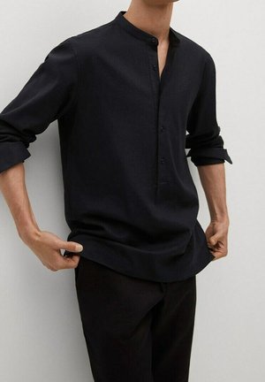 Mężczyzna ubrany w czarną koszulę z długim rękawem i stójką mandaryńską, z podwiniętymi rękawami, trzymający oba brzegi koszuli obiema rękami.