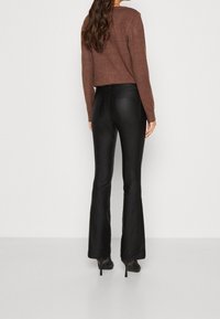 Femme aux longs cheveux bruns portant un pull marron, un pantalon évasé taille haute noir et des chaussures à talons noirs, debout contre un mur blanc.