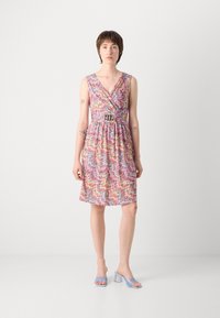 Robe colorée à motif zigzag avec un décolleté en V, taille cintrée et une ceinture décorative. Portée avec des mules à talons transparents bleu clair.