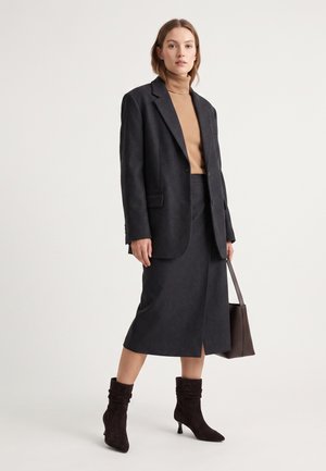 Kvinde iført en mørkegrå blazer og nederdel, tan rullekrave, der holder en brun håndtaske og iført mørke ruskind ankelstøvler med hæle.