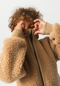 Παιδί με σγουρά μαλλιά τραβάει το φερμουάρ ενός καφέ, χνουδωτού φλις μπουφάν πάνω από το πρόσωπό του σε απλό φόντο.