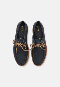 Sapatos de barco de couro azul-marinho com solas de borracha castanha, apresentando um bico arredondado e atacadores bege, exibindo detalhes de costura ao longo das bordas.