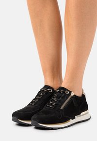 Černé semišové tenisky s kovovými detaily na tkaničkách, bočním zipem a kontrastní bílou a černou podrážkou. Texturované látkové akcenty.