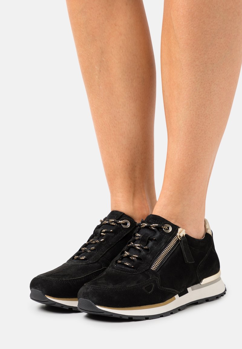Černé semišové tenisky s kovovými detaily na tkaničkách, bočním zipem a kontrastní bílou a černou podrážkou. Texturované látkové akcenty.