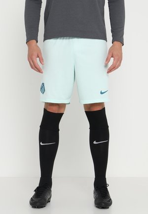 Mężczyzna ubrany w jasnoniebieskie spodenki piłkarskie z logo, czarne wysokie skarpetki oraz czarne korki, stojący na białym tle.