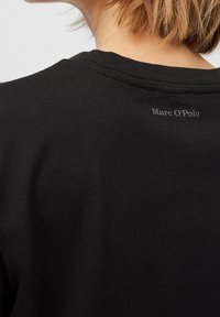 Primer plano del cuello y el hombro de una persona que lleva una camiseta negra de Marc O’Polo con el logo impreso cerca del escote en la parte posterior.