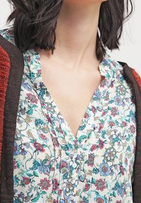 Blouse fleurie présentant des motifs multicolores de fleurs et de feuilles sur un fond clair, avec un devant boutonné et un détail de col.